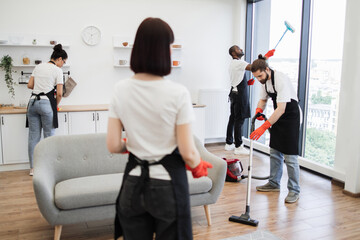 Group of janitors in uniform cleaning office with cleaning equipment. Back view of female boss of...