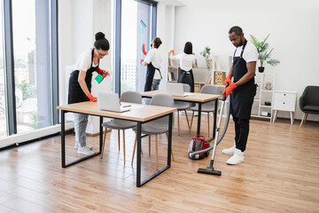 Team of young multicultural cleaners vacuums floor, wipes tables and gadgets, shelves, washes...