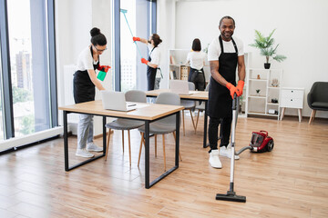 Young adult African American man wearing black apron and red rubber gloves vacuuming floor of modern light office. Professional service team of cleaning company.