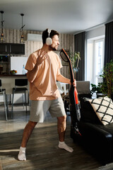 Young man in casualwear and headphones listening to music and singing in electric vacuum cleaner...
