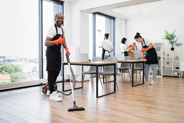 Professional service team of cleaning company. Young adult African American man wearing black apron and red rubber gloves vacuuming floor of modern light office showing thumb up.