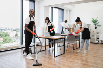 Fototapeta na wymiar Team of multicultural cleaners wipes tables and gadgets, shelves, washes windows in spacious, bright, modern office. Side view of African American man using vacuum cleaner to clean floor from dirt.