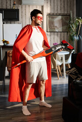 Fototapeta na wymiar Young man in red mantle and mask of superhero holding electric vacuum cleaner while standing in the center of living room at home