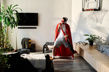 Young man in red superhero mantle singing in vacuum cleaner while standing in the center of bedroom...