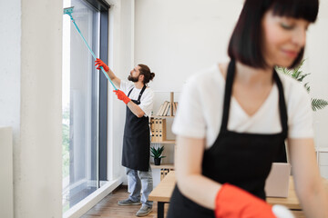 Professional team of people cleaning service in black aprons and red gloves cleans tables, floor, and panoramic windows of spacious studio . Multinational group cleans bright office.