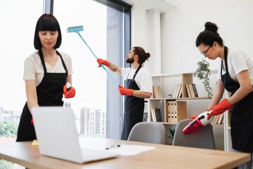 Professional team of people cleaning service in black aprons and red gloves cleans tables, floor, and panoramic windows of spacious studio . Multinational group cleans bright office.