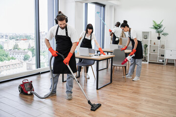 Team of young multicultural cleaners vacuums floor, wipes tables with gadgets, shelves, washes...