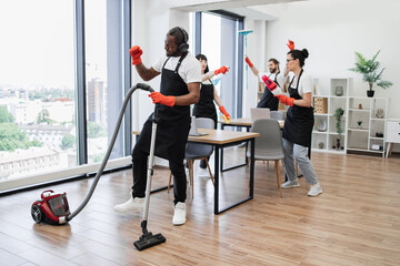 Adult bearded African man in headphones with modern vacuum cleaner and three multiracial cleaners in uniform listening music and dancing with equipment for cleaning on background of modern office.