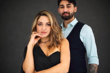 Stylish young ethnic couple embracing against dark gray wall background looking to camera