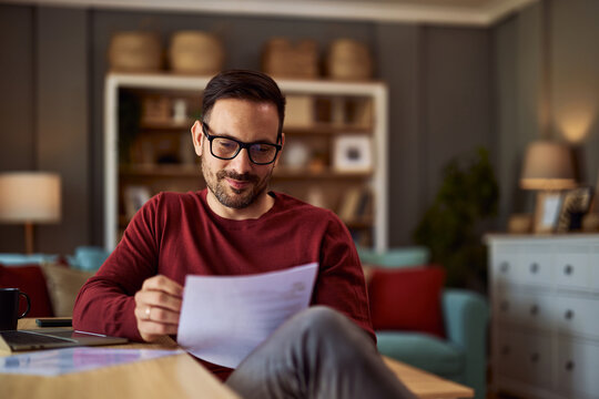 A content male writer revising his work on a paper that he is holding while sitting in his home office.