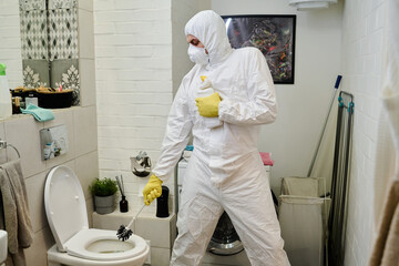 Guy in rubber gloves, respirator and hazmat suit holding brush and plastic bottle of detergent...