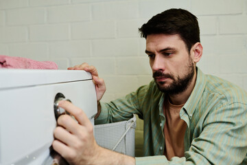 Serious young man squatting by automatic washing machine in bathroom and choosing settings before...