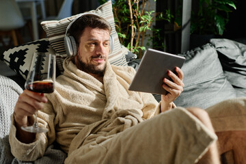 Relaxed young man with glass of wine looking at tablet screen while lying on couch and watching online movie or communicating in video chat