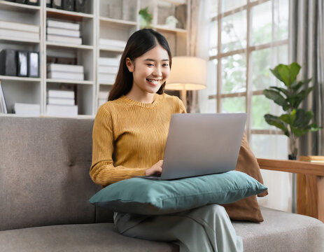 Happy young asian woman using laptop computer while seated on sofa in the living room at home. weekend, watching movie, shopping online.