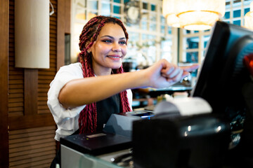 Horizontal photo of a dark-skinned waitress in uniform working in a cafe. An African woman with braided hair is taking out a bill on the touch computer in the restaurant. - obrazy, fototapety, plakaty