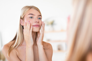 Pretty blonde woman applying moisturizer to face touching cheeks indoors