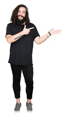 Young man with long hair and beard wearing heavy metal black outfit amazed and smiling to the camera while presenting with hand and pointing with finger.