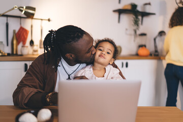 Black father kissing little child while using laptop in kitchen