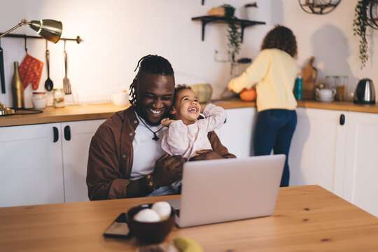 Cheerful ethnic father and cute daughter sitting at dining table in kitchen