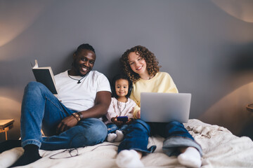 Diverse happy family using laptop while sitting with book on bed