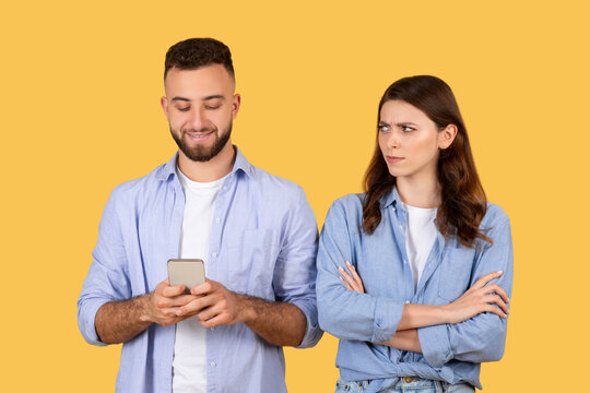 Man smiling at phone with woman looking on skeptically, yellow backdrop
