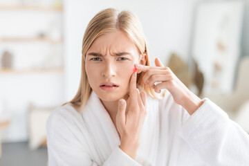 Unhappy blonde young lady squeezing pimples at home bathroom interior