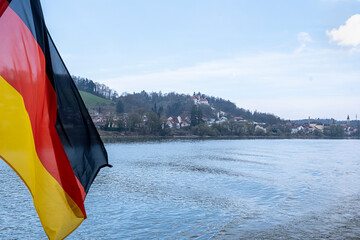 In the foreground the German flag flies in the wind as the ship sails in the foreground. Passau city in the background