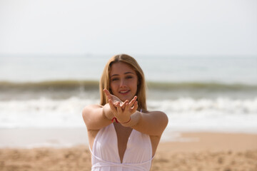 Fototapeta na wymiar Young beautiful blonde woman in white dress is walking on the sand on the shore of the beach on a sunny day. The woman shows her hands clasped together in front. In the background the blue sea.