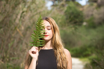 Portrait of a young, beautiful blonde woman dressed in black, half of her face is covered with a green leaf in the forest. The woman is at peace with herself.