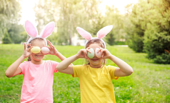 Two happy girls with Easter bunny ears are holding Easter eggs covering their eyes. Children celebrate Happy Easter. Bright sunny day outdoors. The sisters stand nearby in bunny costumes.
 - Powered by Adobe