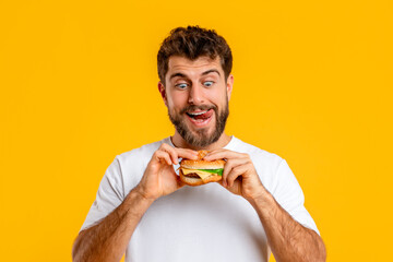 Joyful Hungry Young Guy Eating Burger Posing On Yellow Background