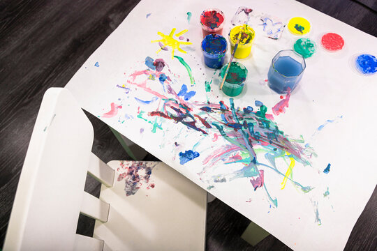 Kids watercolors and children's drawings on white paper, lie on a small kigs table, next to a chair. Kindergarten