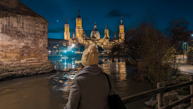 Aragonese Nocturnality: Radiant woman immersed in the grandeur of Zaragoza's Basilica del Pilar, Ebro River, and Stone Bridge. Dazzling reflections manifest admiration in the night.

