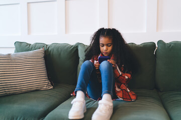 Serious black girl using smartphone while sitting on sofa