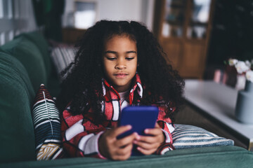 Young African-American girl engaged with her handheld smartphone device, comfortably snuggled on a...