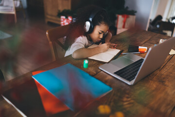 Focused little girl in headphones using laptop while doing homework at table