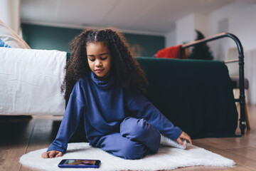 Contemplative young African-American girl sitting on the floor at home with a smartphone by her...