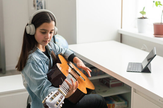 Latin girl with headphones listening to her online music lessons. Artistic kid playing the acoustic guitar and learning the chords