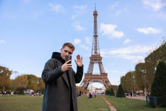 A man in a green coat near the Eiffel Tower in Paris makes a video call and takes a happy selfie. Capturing the essence of holidays and travel in France, it emphasizes connection and internet networks