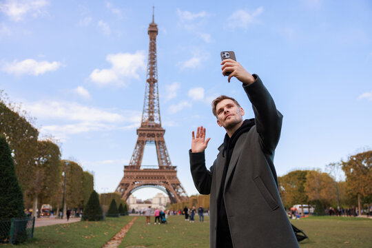 Man near Eiffel tower in Paris making video call from cell phone. Happy Tourist taking selfie. Male in green coat. Holidays and travel concept in France. Connection and internet network