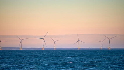 Wind farm field in the sea near the coast, during sunset with mountains and clear, colorful sky in the background.