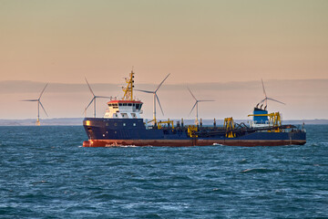 Dredger vessel working in the sea, with wind turbines on the background on a sunny day.
