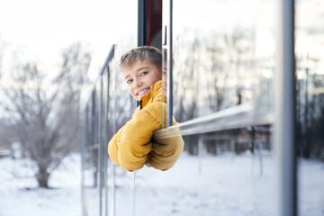 Front view of funny child male smiling looking at camera leaning out bus window. Crop of cheerful young guy wearing orange winter jacket travelling by public transport. Concept of holiday trip.