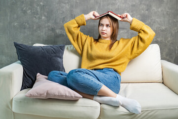 young woman is sitting on the sofa, holding a book over her head with her hands