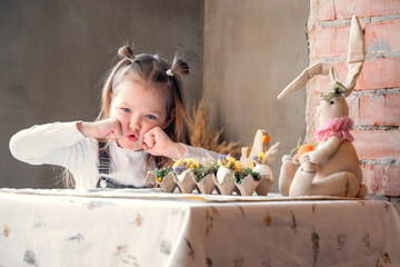 Cute and funny kid. Girl sitting at kitchen table, coffee table, little lady. Easter eggs, Easter hare, wooden chicken, preparation for Easter, flowers