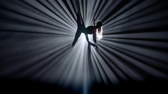 Silhouette of female isolated on black background bright spotlight. Girl aerial dancer performing balancing split on air silk.