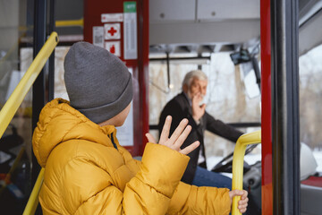 Back view of little boy in grey cap standing near open door saying goodbye to driver in transport. Crop of child male in orange jacket waving to driver getting off bus. Concept of politeness.