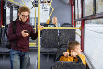 Fototapeta na wymiar Front view of happy adult male using phone smiling standing in moving bus. Crop of cute young man in hoodie travelling with blur blonde and little boy sitting in urban transport. Concept of city life.