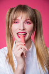 Naklejka premium Portrait of a young woman with braces and bright makeup chewing gum on a pink background. Vertical photo.