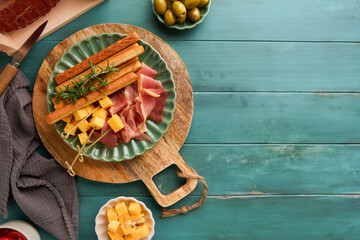 Slices of prosciutto or jamon. Antipasto Delicious grissini sticks with prosciutto, cheese, rosemary, olives on green plate on old wooden blue background. Appetizers table with italian snacks Top view